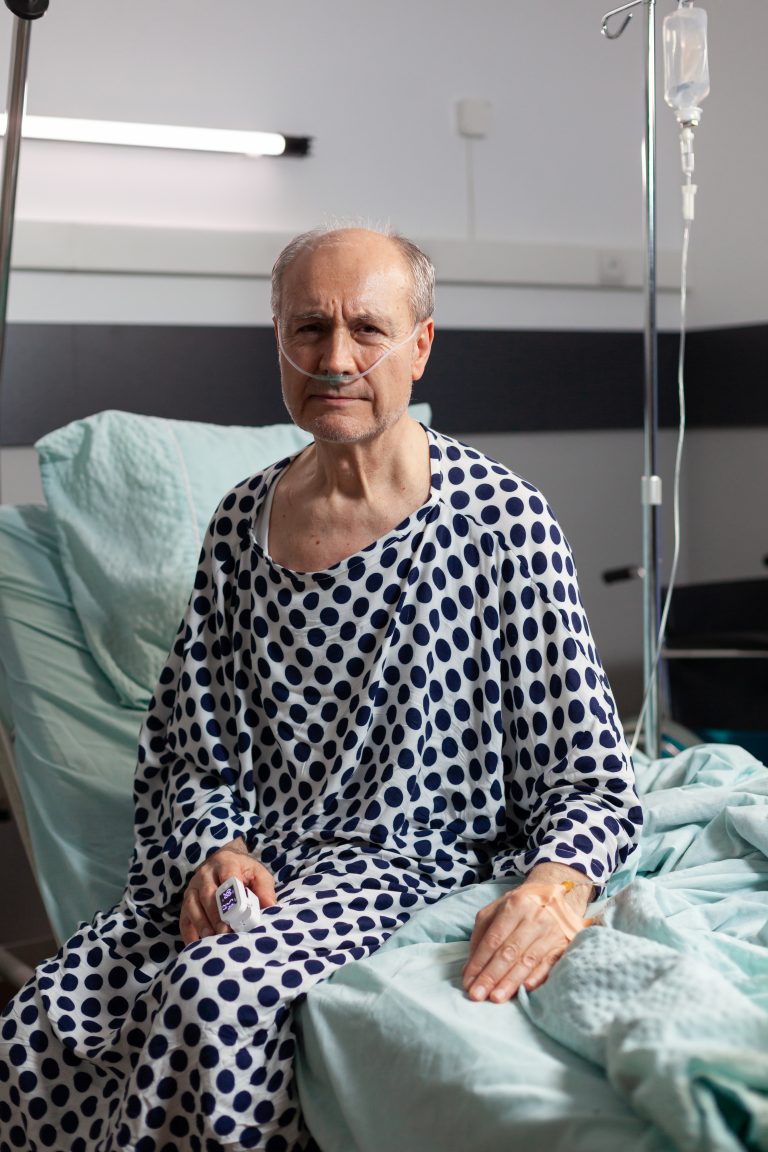 Portrait of sad, unwell senior man sitting on the edge of hospital bed